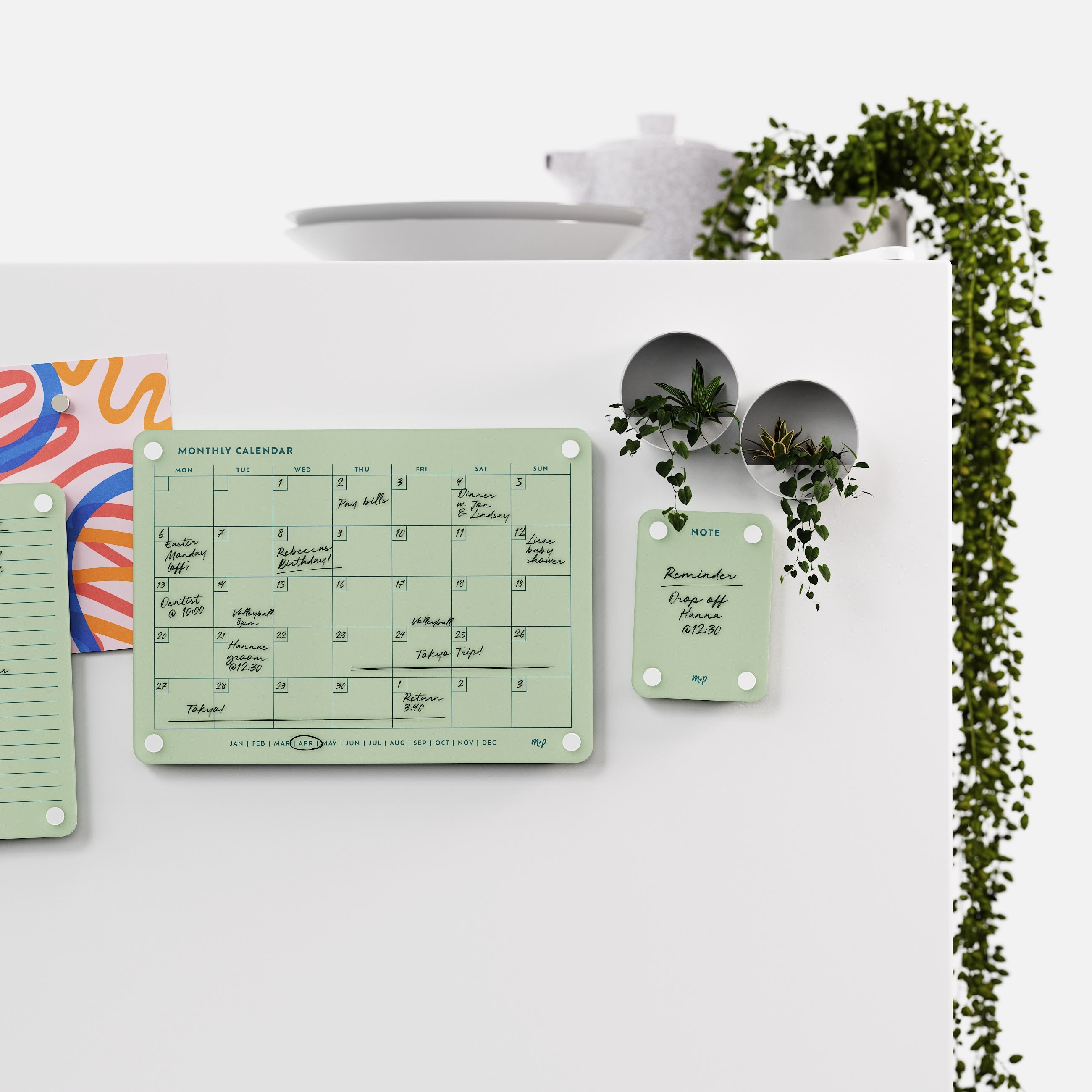 Matcha green monthly calendar and note board on a white kitchen wall with magnetic planters and trailing ivy